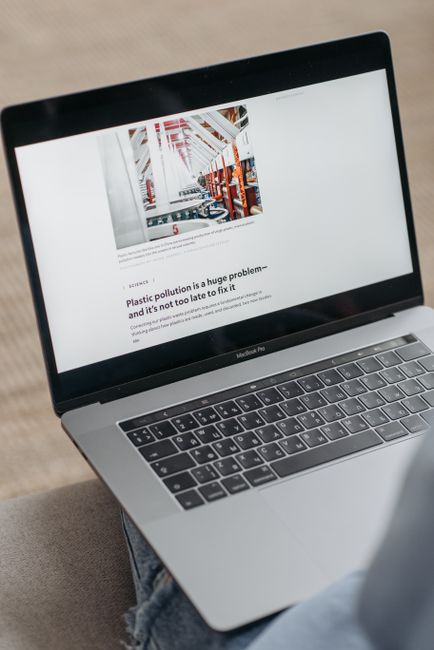 A silver MacBook Pro laptop resting on a person's lap, displaying a webpage titled 'Plastic pollution is a huge problem - and it's not too late to fix it,' with an image of a warehouse interior featur